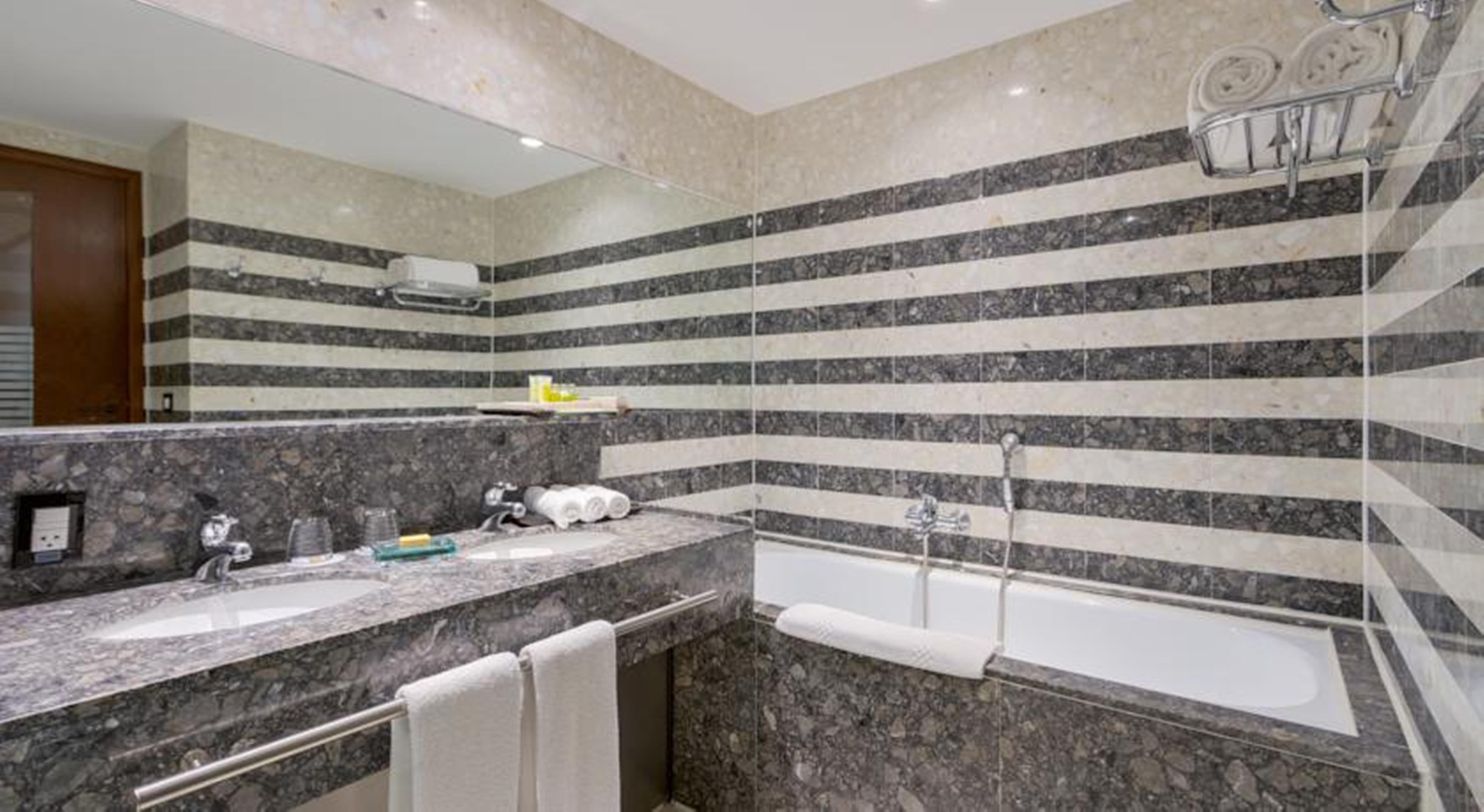 Royal Tulip Brasilia Alvorada Hotel ภายนอก รูปภาพ The photo shows a modern bathroom featuring a combination of marble and stone finishes. The walls are decorated with horizontal stripes in various shades of gray and cream. There is a bathtub with a white exterior, and a showerhead is mounted on the 