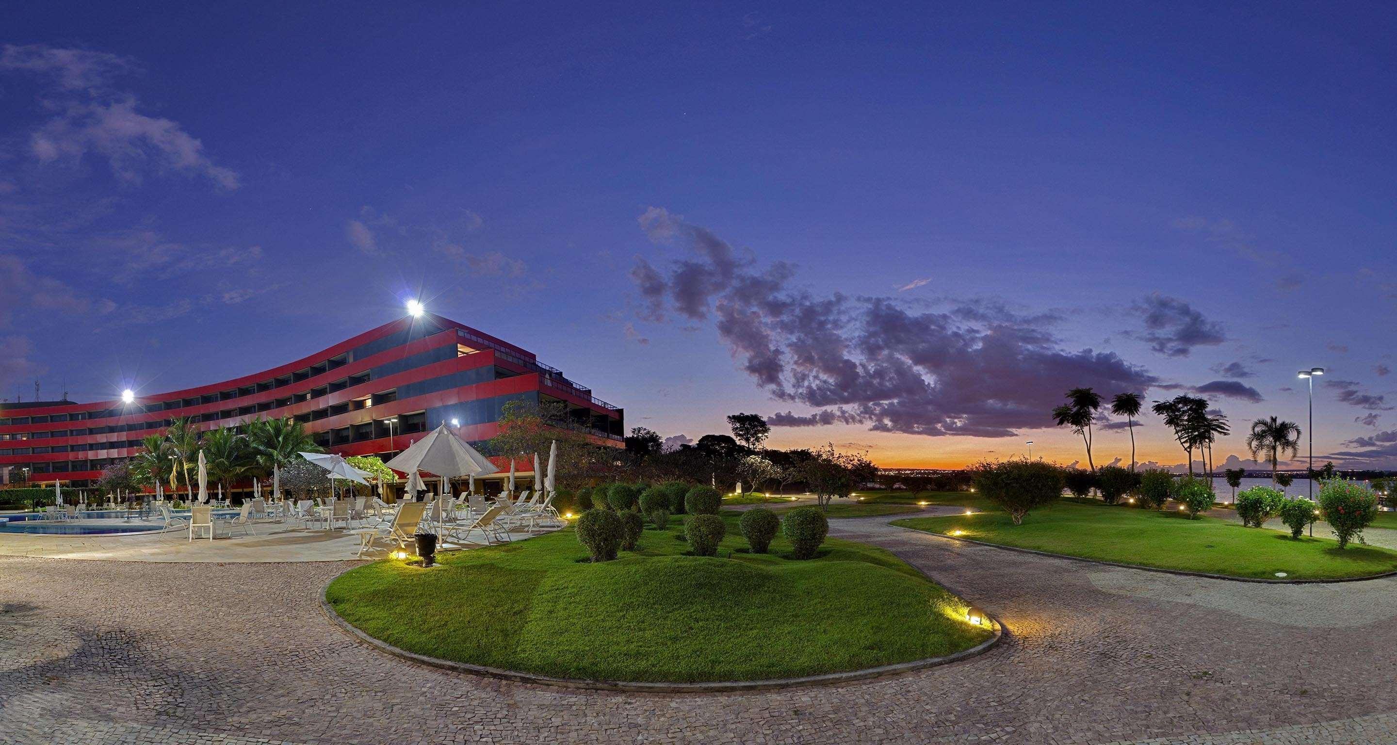 Royal Tulip Brasilia Alvorada Hotel ภายนอก รูปภาพ The photo depicts a modern building, possibly a hotel or resort, with a striking red exterior. It is set in a landscaped area that features well-maintained greenery and ornamental plants. In the foreground, there are lounge areas with umbrellas, sugg