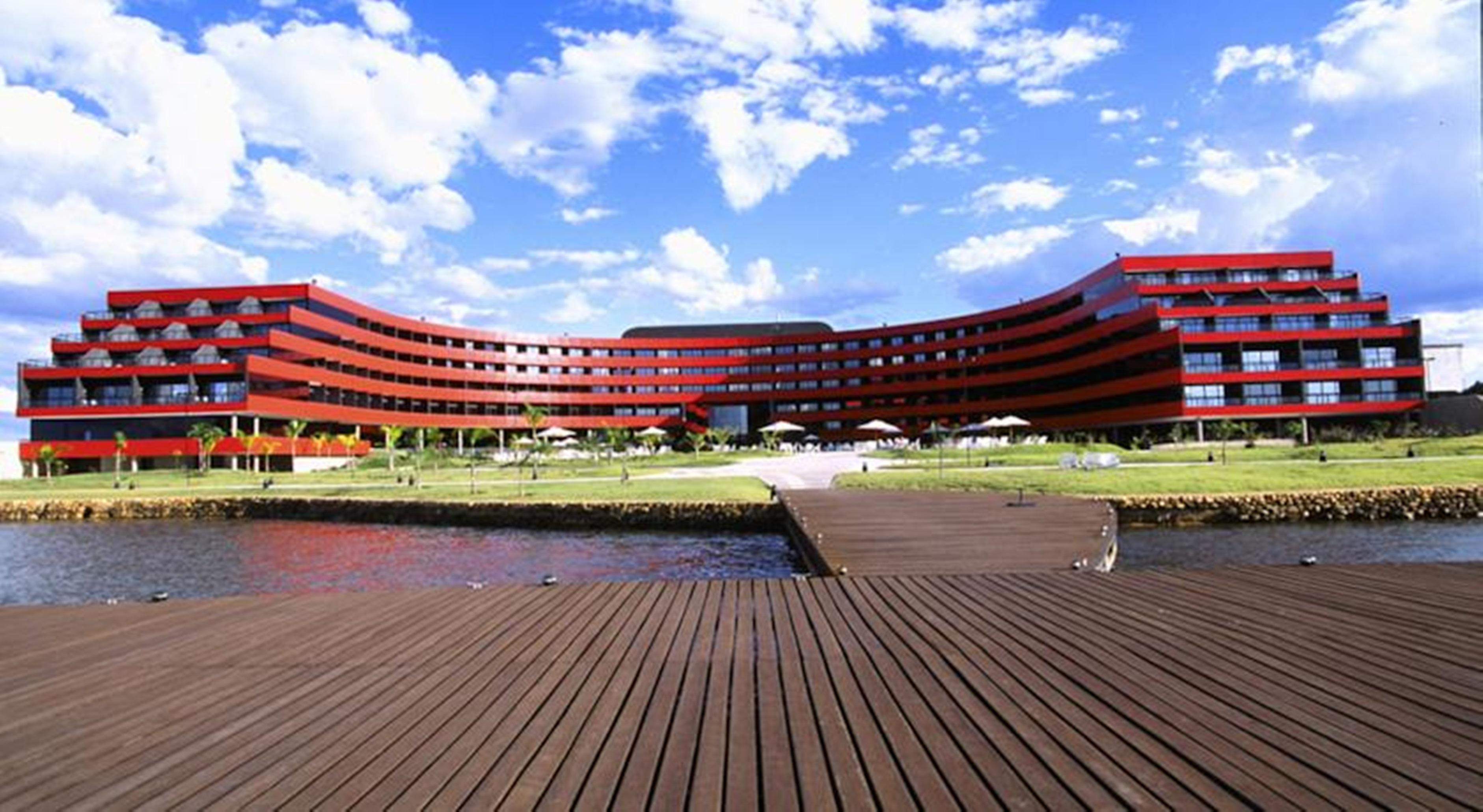 Royal Tulip Brasilia Alvorada Hotel ภายนอก รูปภาพ The image shows a modern, curved building with a striking red facade, situated near a body of water. The architecture features multiple levels with large windows that create a sleek and contemporary appearance. In front of the building, there is a wo