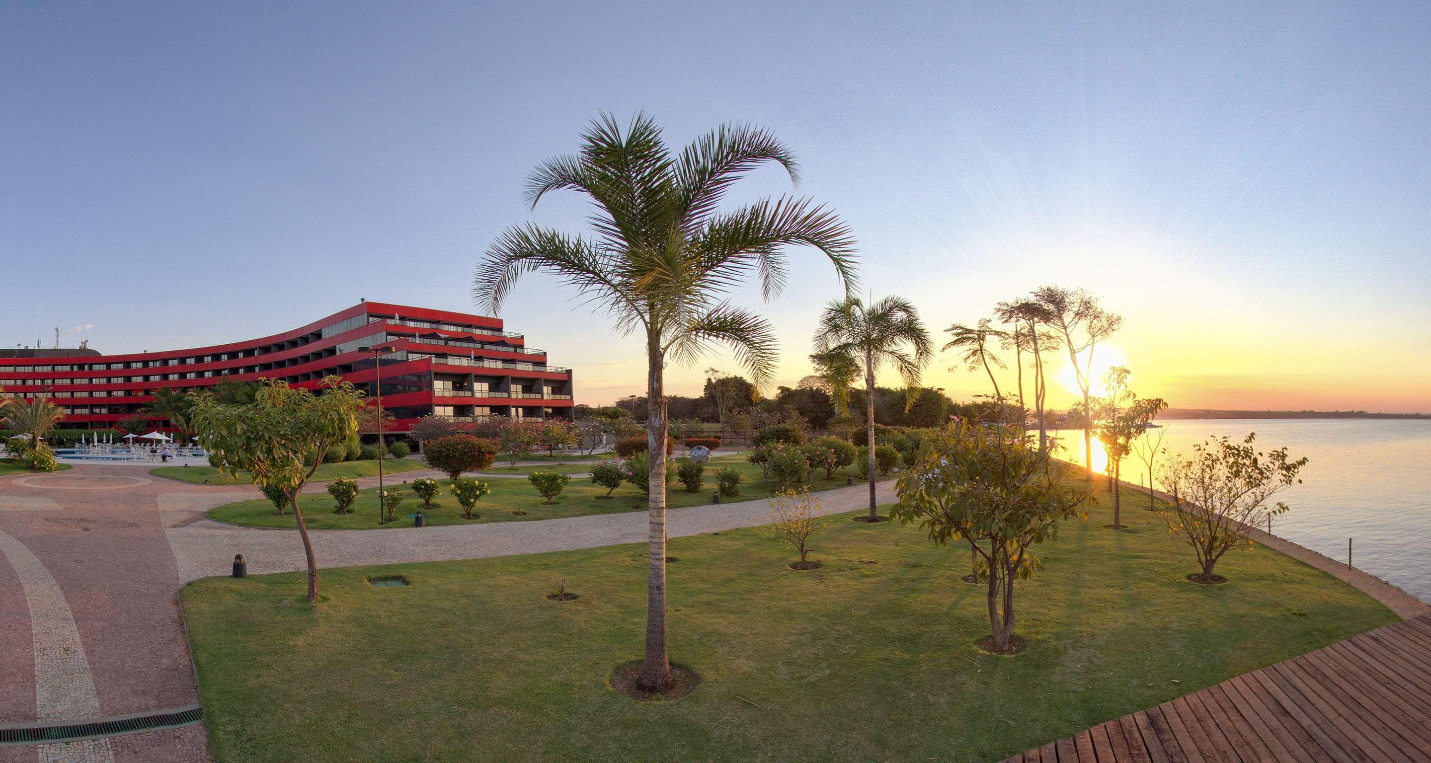 Royal Tulip Brasilia Alvorada Hotel ภายนอก รูปภาพ The photo depicts a scenic view featuring a red multi-story building alongside a serene waterfront. In the foreground, there are several palm trees and lush green lawns, creating a tropical atmosphere. The sun is setting in the background, casting wa