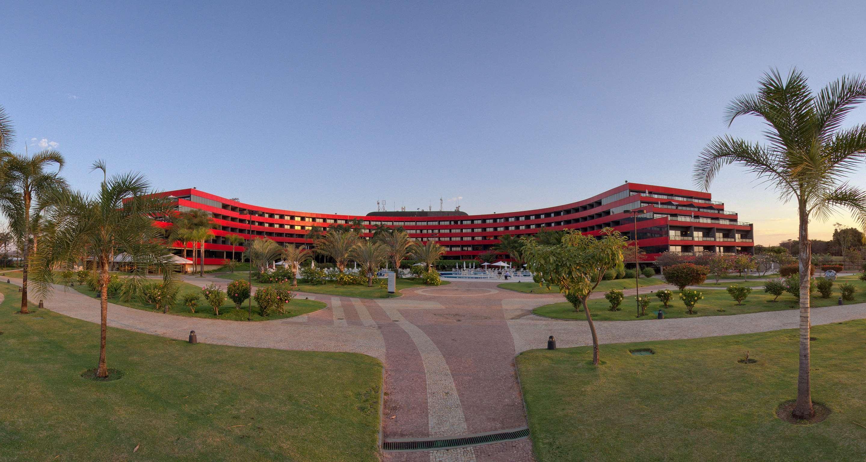 Royal Tulip Brasilia Alvorada Hotel ภายนอก รูปภาพ The photo shows a large, curved building with a vibrant red exterior. It appears to be a hotel or resort, surrounded by well-maintained gardens and green spaces. The landscape includes manicured lawns, small trees, and pathways, suggesting a pleasant