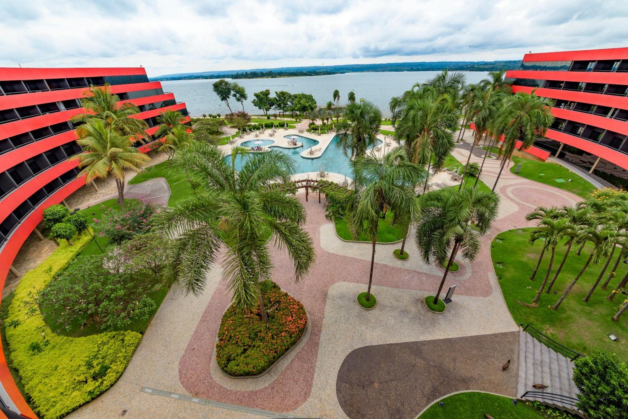 Royal Tulip Brasilia Alvorada Hotel ภายนอก รูปภาพ The photo shows a scenic view of a courtyard area in a residential complex. There are multiple red buildings surrounding a pool area, which is surrounded by palm trees and landscaped gardens. The pool features a curvy design, and there are pathways m