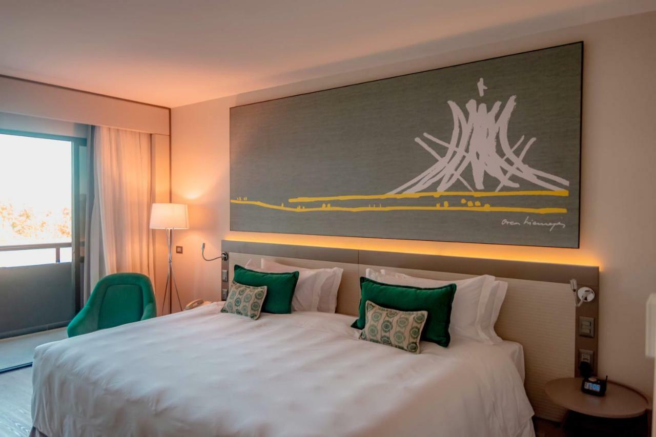 Royal Tulip Brasilia Alvorada Hotel ภายนอก รูปภาพ The photo shows a modern hotel room featuring a large bed with white bedding and two green accent pillows. Above the bed, there is a large piece of artwork depicting a stylized mountain or volcanic shape with yellow accents. The room has a contempora
